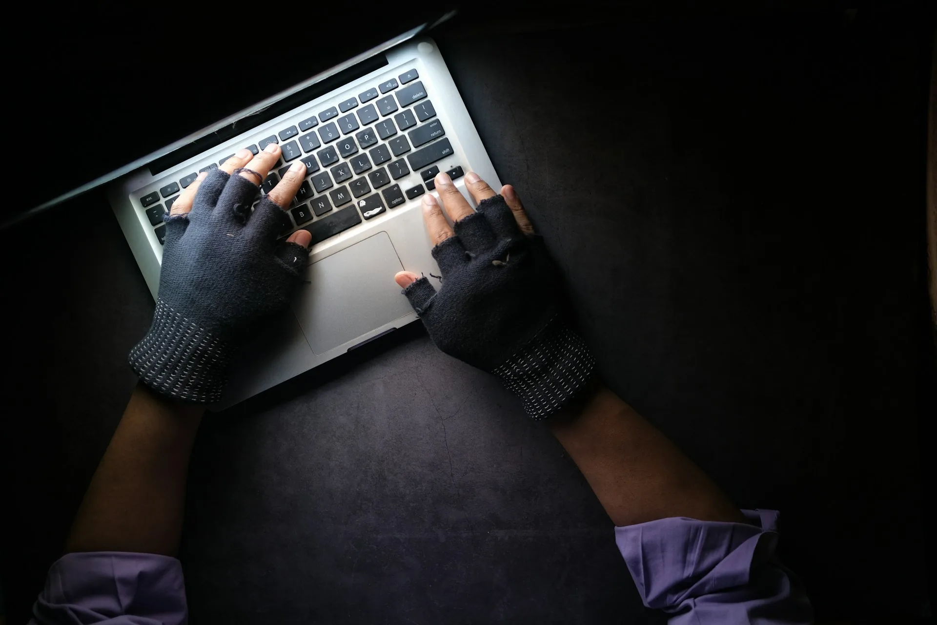 Overhead view of hands in fingerless gloves typing on a laptop in a dark room