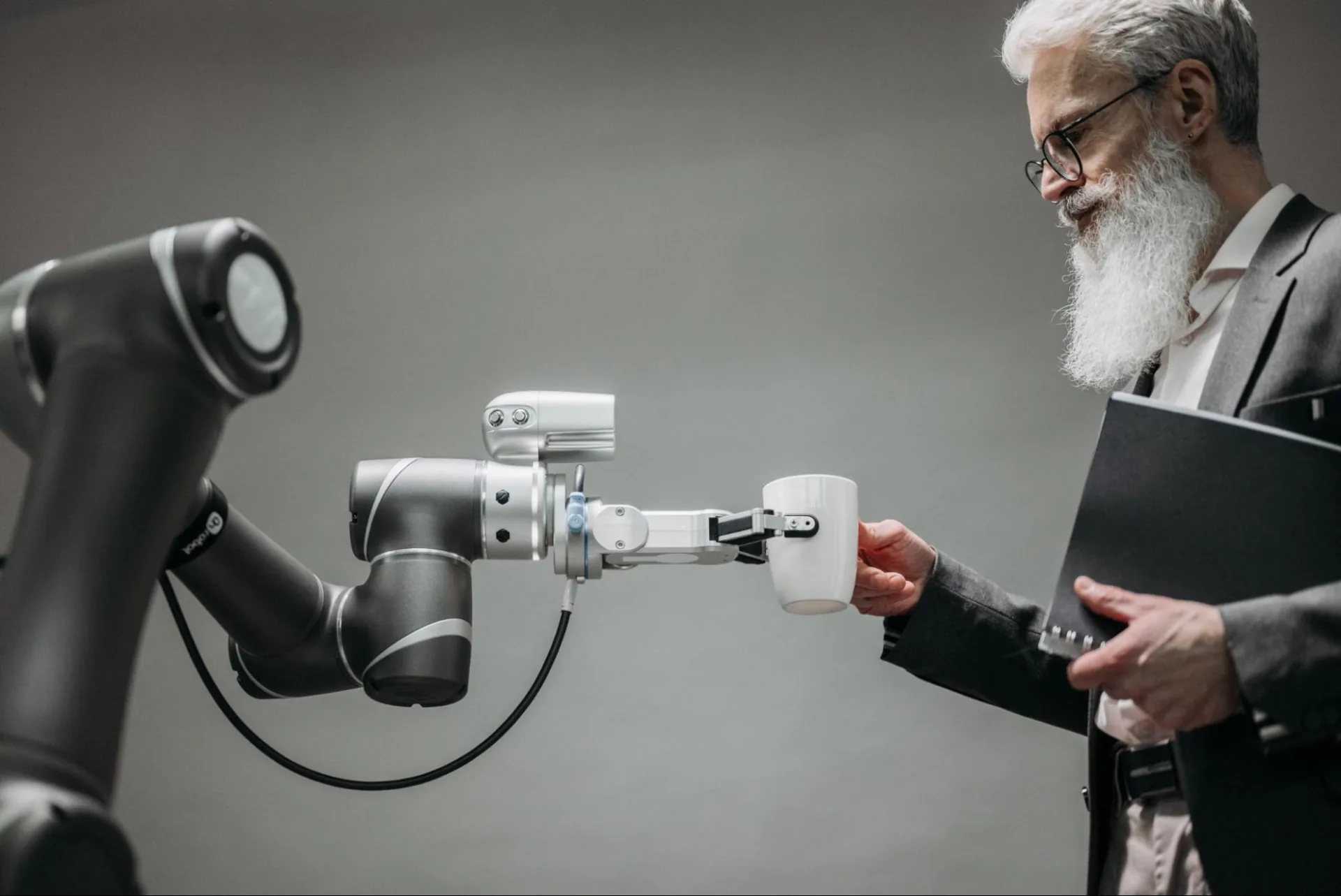 A gray robotic arm passing a white coffee mug to a gray-bearded man in glasses and a suit, who holds a black folder against a plain gray backdrop