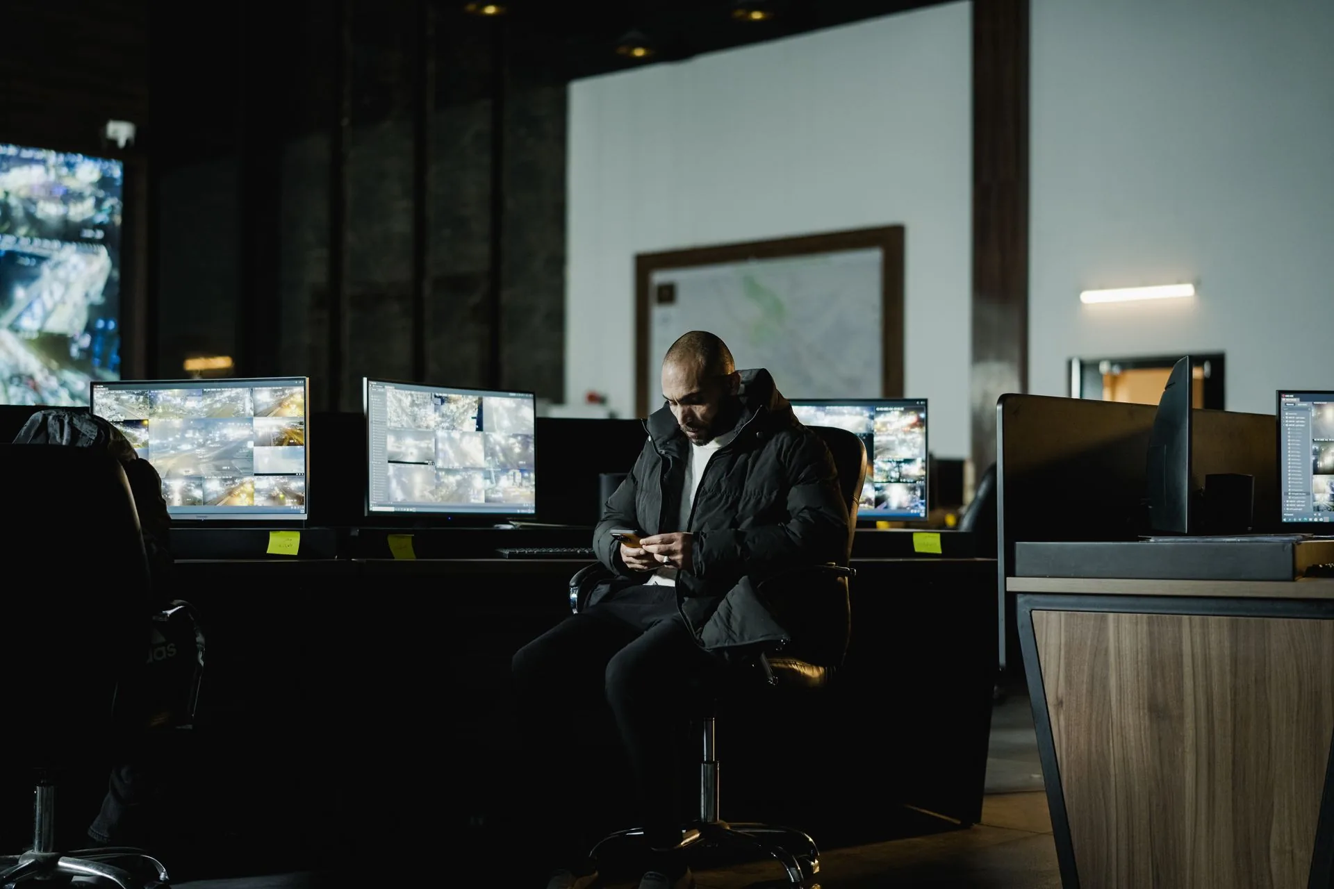 A security operator checking a phone in a dim control room, surrounded by monitors showing grids of live city camera feeds