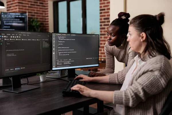 Two developers at a shared desk looking at code on a monitor in a brick-walled office