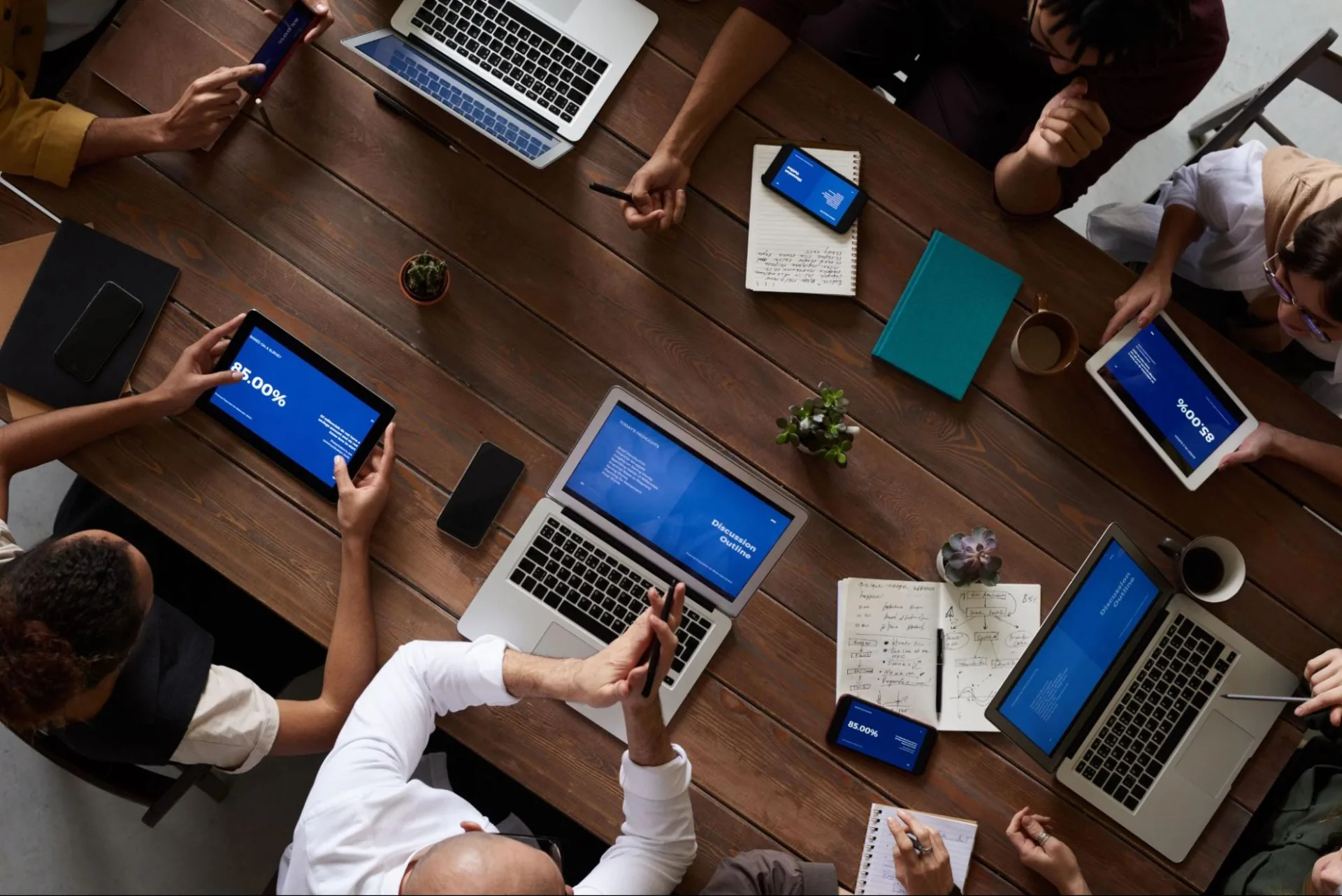 Overhead view of a team seated around a wooden table covered in laptops, tablets and phones showing blue discussion-outline slides, alongside notebooks and coffee cups